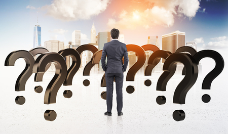 Rear view of a businessman standing with his arms in pockets surrounded by questions marks. There is a cityscape in the background.の写真素材