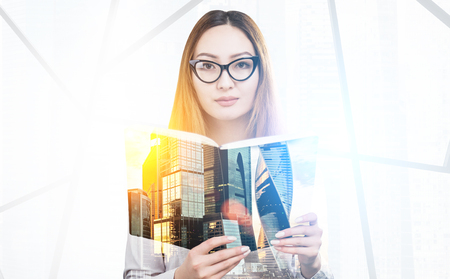 Portrait of a young woman wearing glasses and holding a book and standing in an office. There is a cityscape in the foreground. Toned image. Double exposureの写真素材