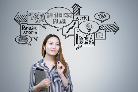Portrait of an Asian businesswoman holding a tablet computer and standing near a gray wall with business speech bubbles drawn on it.の写真素材