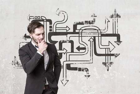 Portrait of a pensive bearded businessman in a suit thinking near a concrete wall with tangled arrows on it.の写真素材