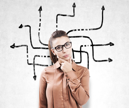 Portrait of a woman in glasses and a brown blouse standing near a concrete wall with an arrow maze on it.の写真素材