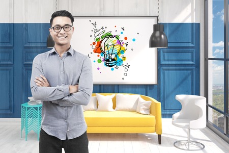 Portrait of a happy Asian businessman wearing glasses standing in an office with a small light bulb sketch on a whiteboard.の写真素材