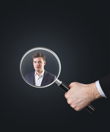 Close up of a hand of a businessman holding a giant magnifying glass with a portrait of a man inside it. Concept of recruitment, Mock up.の写真素材