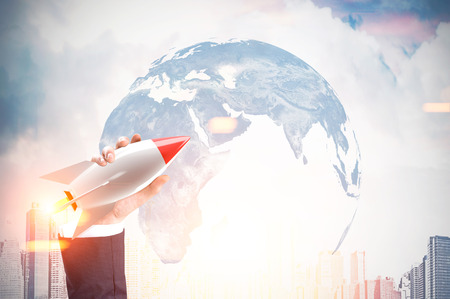 Close up of a hand of a businessman holding a toy rocket and the Earth image and city in the backgrounToned image. Double exposureの写真素材