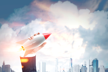 Close up of a hand of a businessman holding a toy rocket and a clowdy cityscape in the background. Toned image. Double exposureの写真素材
