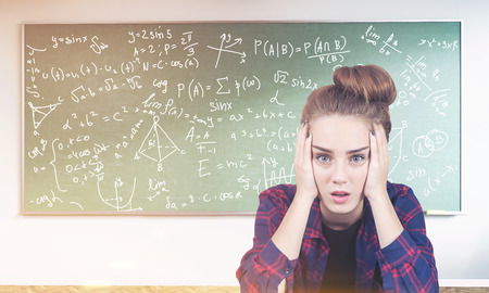 Portrait of a stressed out teen girl sitting near a green chalkboard with formulas written on it. Concept of exact sciences. Toned imageの写真素材