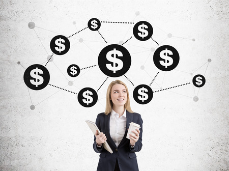 Portrait of a smilng blond businesswoman standing with a cup of coffee and a clipboard near a concrete wall with a money network sketch on it.の写真素材