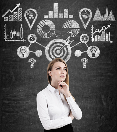 Portrait of a young blond pensive businesswoman standing near a blackboard with a target sketch drawn on it.の写真素材