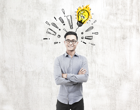 Portrait of a happy Asian businessman standing near a concrete wall with a ligth bulb surrounded by exclamation marks on it.の写真素材