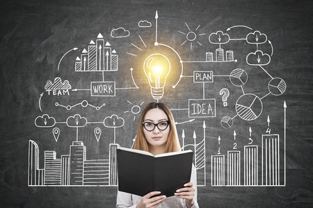 Portrait of an Asian businesswoman wearing glasses and reading a book while standing near ablackboard with a business idea drawing.の写真素材
