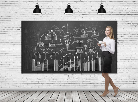 Side view of a blond businesswoman wearing a white blouse and a black skirt and holding her cup of coffee while standing near a blackboard with a business scheme drawn on it.の写真素材