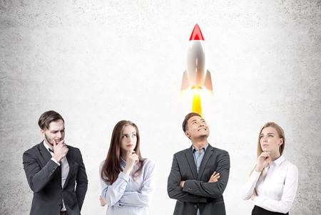 Portrait of a group of businessmen and businesswomen standing near a concrete wall brainstorming. A rocket is taking off above one of themの写真素材