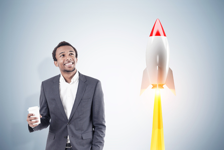 Portrait of an African American businessman holding his paper cup of coffee and standing near a gray wall with a rocket taking off.の写真素材