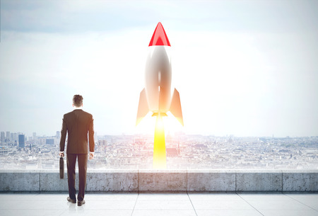 Rear view of a businessman with a suitcase standing on a roof of a tall building and looking at a red and white rocket taking off in a city. Toned imageの写真素材