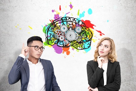 Portrait of an Asain businessman and a blond woman standing near a concrete wall with a colorful brain sketch and cogs drawn on it.の写真素材