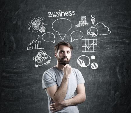 Portrait of a bearded man wearing a gray T-shirt and standing near a blackboard with a business bubble sketch drawn on it.の写真素材