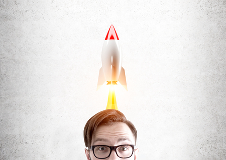 Close up of a head of a man wearing glasses and standing near a concrete wall with a rocket flying above his head. Concept of a start upの写真素材