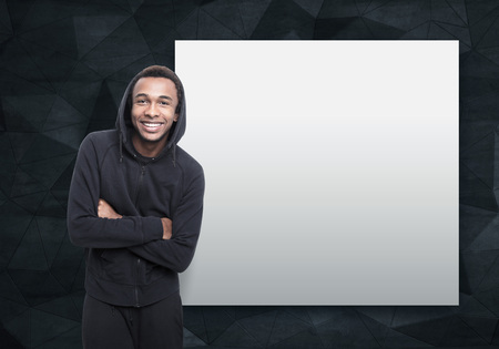 Portrait of a smiling African American young man wearing a black hoodie and standing with crossed arms near a blank poster. Mock upの写真素材