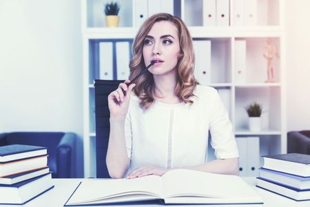Portrait of a pensive red haired businesswoman at her workplace in a white office. She is holding a pen and sitting in front of an open notebook. Toned image.の写真素材