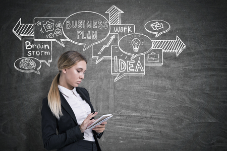 Side view of a businesswoman with a notebook writing near a blackboard with a business plan sketch drawn on it.の写真素材