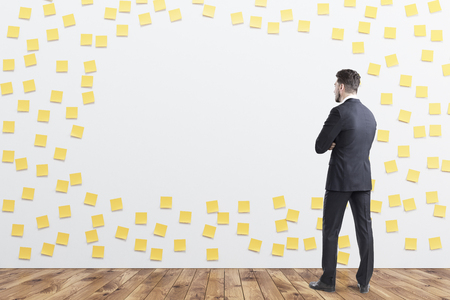 Rear view of a businessman in gray looking at a wall covered by yellow sticky notes and a blank piece. 3d rendering, mock upの写真素材