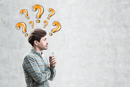Side view of a young man wearing a green shirt and standing near a concrete wall with orange question marks drawn above his head. Mock upの写真素材