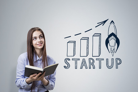 Portrait of a smiling woman in a blue shirt holding a book and standing near a gray wall with a start up graphの写真素材