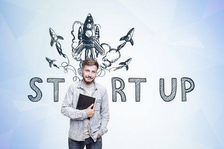 Portrait of a young bearded man holding a black book and standing near a blue wall with a start up sketchの写真素材