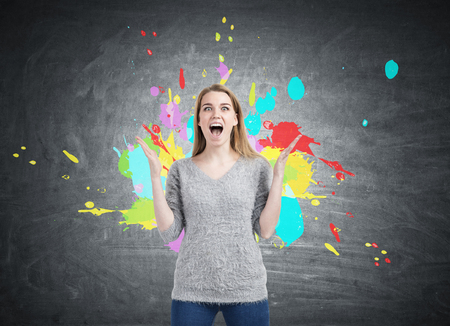 Portrait of a blond woman in a gray sweater screaming with joy standing near a blackboard with color...