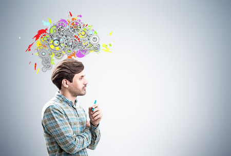 Side view of a young man holding a marker and standing near a gray wall with a colored brain shape and gears on top of it. Mock upの写真素材