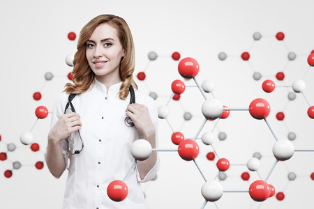 Gorgeous ginger woman doctor in a uniform with a stethoscope is smiling near a white wall with a red and white molecule model on it. Mock upの写真素材