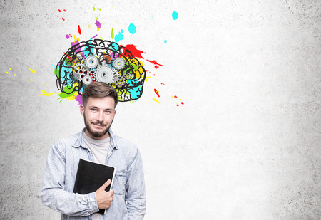 Skeptical young businessman with wavy hair wearing glasses standing near a concrete wall with a colorful brain sketch with gears on top of it. Mock upの写真素材