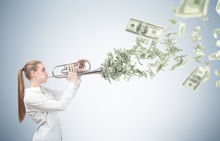 Side view of a blond woman with a trumpet standing near a gray wall playing on her trumpet. Dollar...