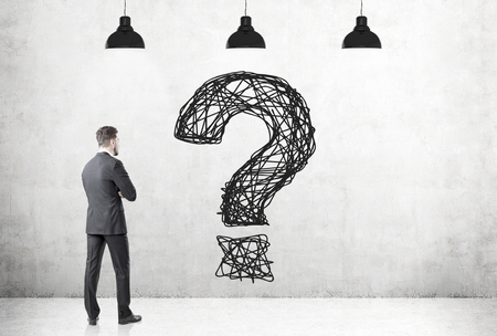 Rear view of a businessman wearing a gray suit looking at a large black question mark drawn with a marker on a concrete wallの写真素材