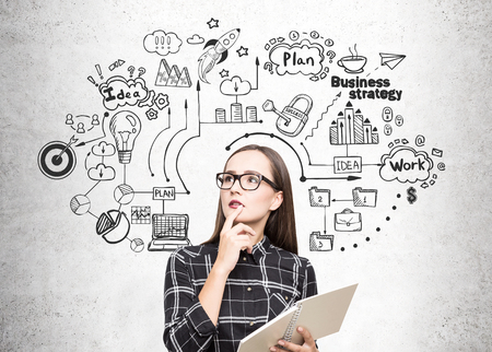 Portrait of a geek girl in glasses standing near a concrete wall with a copybook and a business strategy icons drawn on itの写真素材
