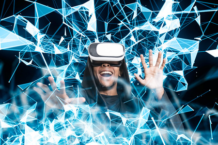 Portrait of an excited African American man wearing a hoodie and VR glasses. He is interacting with...