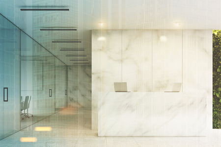 Front view of a white reception desk with two laptops standing on it in front of a marble office wall. There are glass wall offices to the left. 3d rendering, mock up, toned imageの写真素材