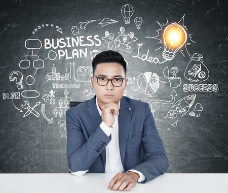 Portrait of a young Asian businessman in glasses sitting near a blackboard with a business plan sketch and a small glowing light bulb on itの写真素材