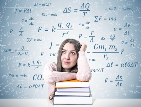 Portrait of a young woman wearing a pink sweater and sitting at a white table with a pile of colorful books. She is dreaming near a gray wall with different formulas written on it. Mock upの写真素材