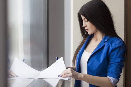 Side view of a woman wearing a blue jacket writing on a sheet of paper on a window sill in an office lobbyの写真素材
