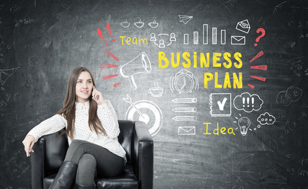 Young businesswoman with fair hair smiling and sitting in a leather armchair and talking on her smartphone. Blackboard with a business plan sketchの写真素材