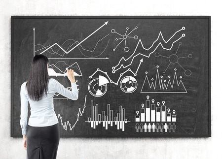 Rear view of a black haired businesswoman wearing a blue shirt and a black skirt drawing a graph on a blackboard.の写真素材