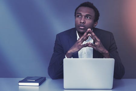 Serious African American businessman is sitting at his table with a laptop and a book. Mock up toned imageの写真素材