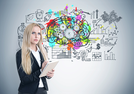 Blonde businesswoman wearing a black suit and a white shirt and holding a copybook is standing near a gray wall with a colorful brain with cogs and a business scheme.の写真素材