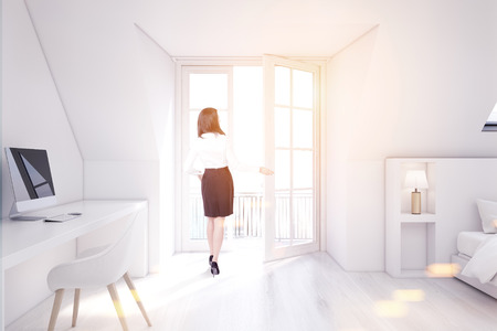 Woman in a white attic bedroom with a home office, white walls and wooden floor, a table with a computer and a bed. 3d rendering mock up toned imageの写真素材