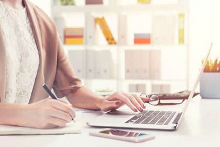 Close up of an unrecognizable young woman wearing a white blouse and a brown short sleeve jacket. She is writing and using her laptop. Toned imageの写真素材