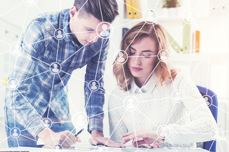 Beautiful businesswoman is sitting at a table in a white office. Her colleague is standing near her and looking at a document on the desk. Network. Toned image double exposureの写真素材