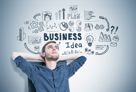 Portrait of a bearded young man sitting in a modern armchair with hands behind his head, looking upwards and thinking. Gray wall background with a business idea sketch on it.の写真素材