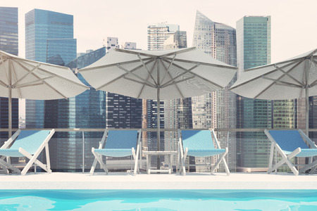 Blue deck chairs are standing under beach umbrellas near a swimming pool. A pale blue cloudless sky and a cityscape. Close up. 3d rendering mock upの写真素材