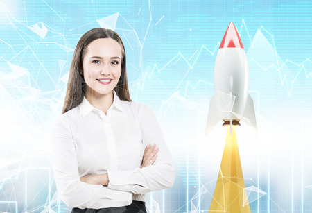 Young and confident businesswoman with crossed arms is standing against a cityscape with a startup rocket and graphs in the sky. Double exposureの写真素材